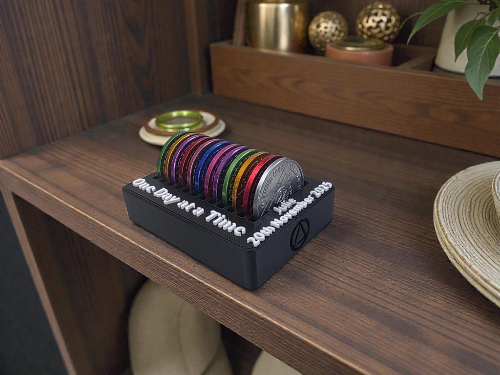 May include: A black coin holder with the text "One Day at a Time" and "Julie 20th November 2025" in white. It holds a variety of colourful coins and a silver coin. The holder sits on a wooden shelf, with other decorative items in the background.