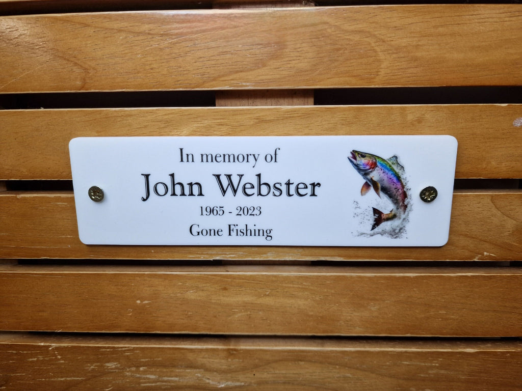 May include: White memorial plaque with black text that reads "In memory of John Webster 1965 - 2023 Gone Fishing". A colourful image of a trout jumping out of the water is on the right side of the plaque.