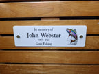 May include: White memorial plaque with black text that reads "In memory of John Webster 1965 - 2023 Gone Fishing". A colourful image of a trout jumping out of the water is on the right side of the plaque.