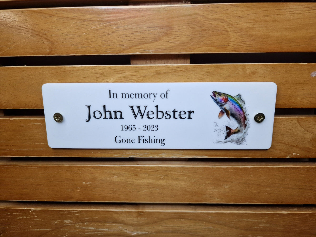 May include: White memorial plaque with black text that reads "In memory of John Webster 1965 - 2023 Gone Fishing". A colourful image of a trout jumping out of the water is on the right side of the plaque.