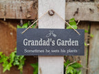 May include: A black slate garden sign with white text that reads "Grandad's Garden Sometimes he wets his plants". The sign is hanging on a wooden fence post with twine.