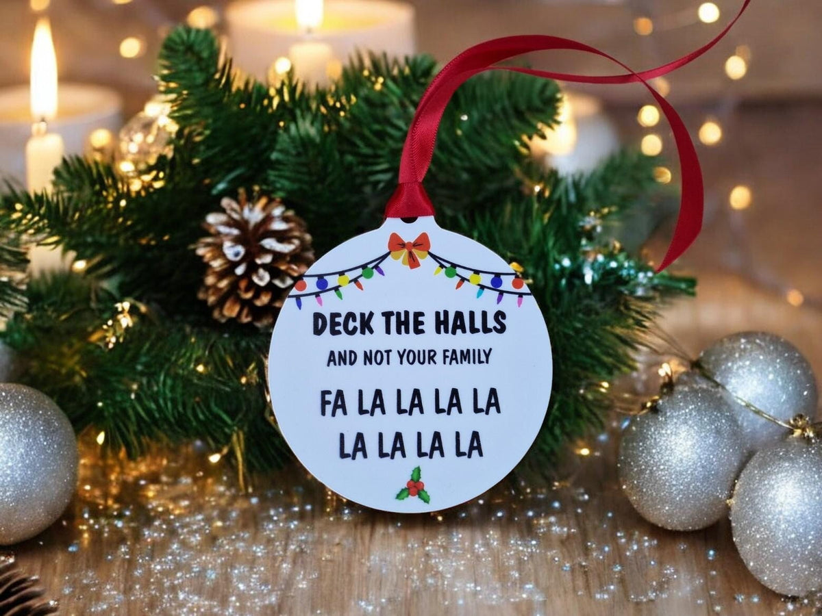 May include: White round Christmas bauble with the text "Deck the Halls and not your family" and "Fa La La La La La La La La" in black text. The bauble is decorated with a red bow and colourful lights.