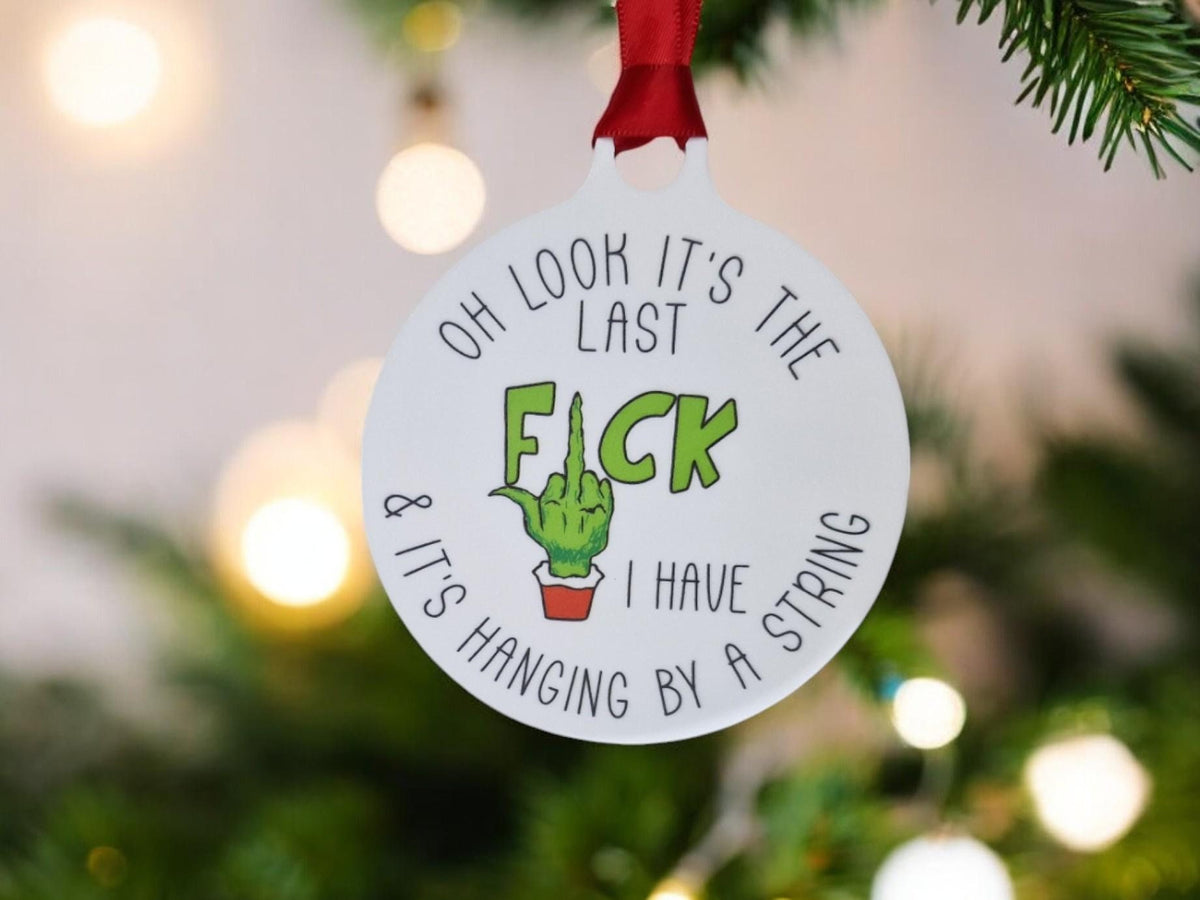 May include: White round Christmas bauble with the text "Oh look it's the last F**k & it's hanging by a string I have". A green hand with a middle finger is shown in the centre of the bauble.